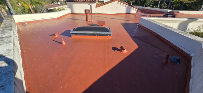 Flat red roof with small skylight and multiple vent pipes on a sunny day, surrounding white parapets and palm trees in the background.